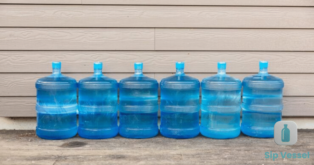 Six large blue plastic bottles are neatly lined up against a wall, showcasing their uniform size and color.